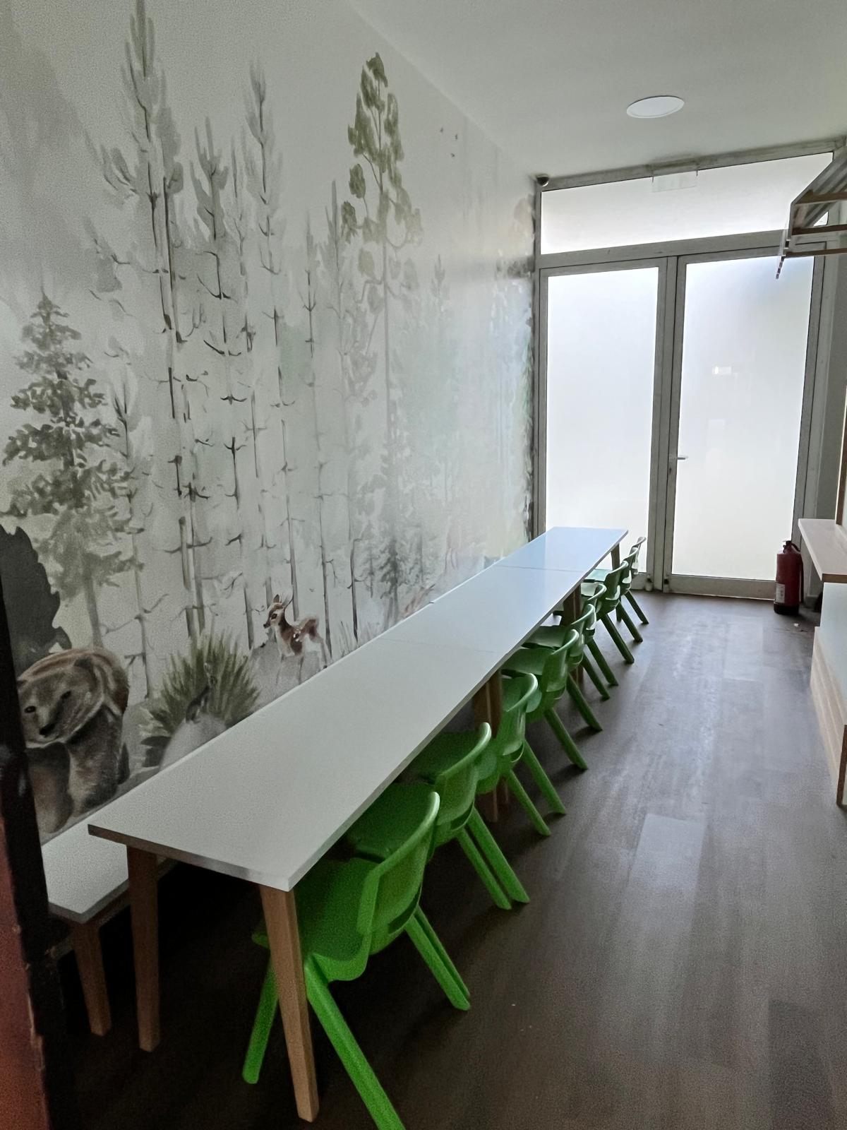 Mesa corrida con sillas verdes frente a un mural de bosque con un cervatillo