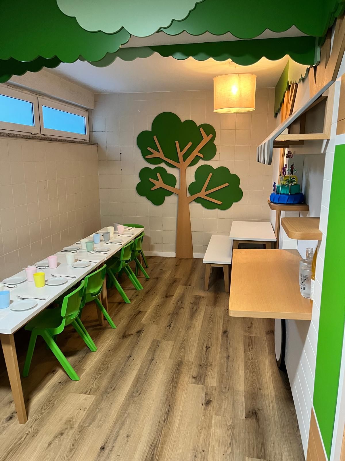 Mesa larga preparada para un cumpleaños con sillas verdes y un árbol recortado en la pared